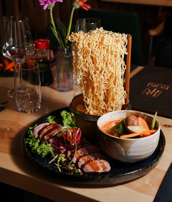 Ramen mit hochgehobenen Nudeln, Suppe und Beilagen am Tisch.